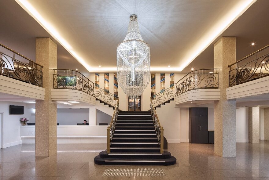 The marble interior of the Victoria Hotel in Melbourne, with a central marble staircase in a double height atrium with ornate iron balustrades and a large chandelier.