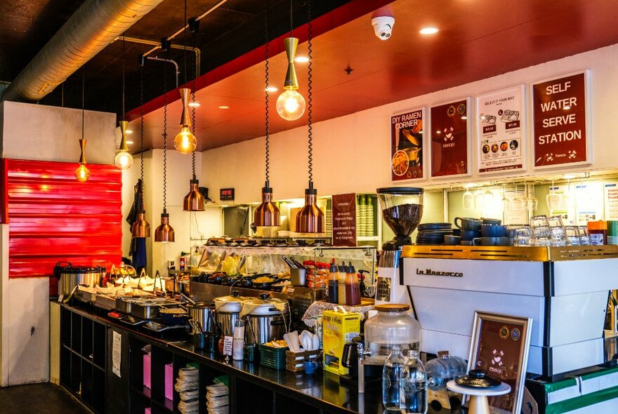 Cafe interior with food in trays and coffee machine, pendant lighting and crowded counters.