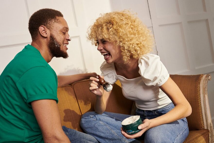 A laughing woman holding an open tub of face cream in her hands, seated on a couch opposite a man with closed eyes.