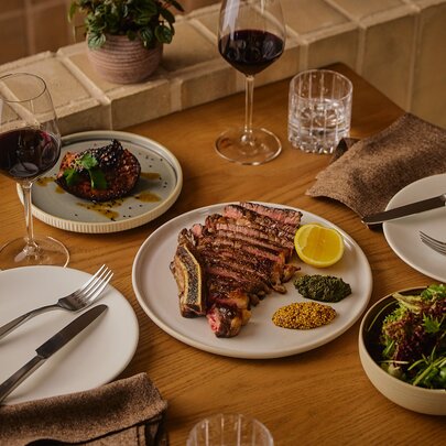 A wooden table set with two place settings, with a sliced rib eye and condiments plus side dishes and wine.