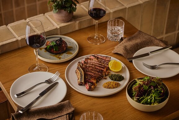 A wooden table set with two place settings, with a sliced rib eye and condiments plus side dishes and wine.