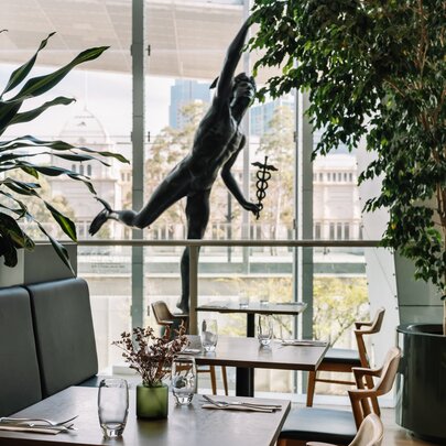 Restaurant interior with tables set for service in the foreground, a bronze sculpture in the open space behind the tables, and through the large windows a view of an ornate Victorian building and trees in the distance.
