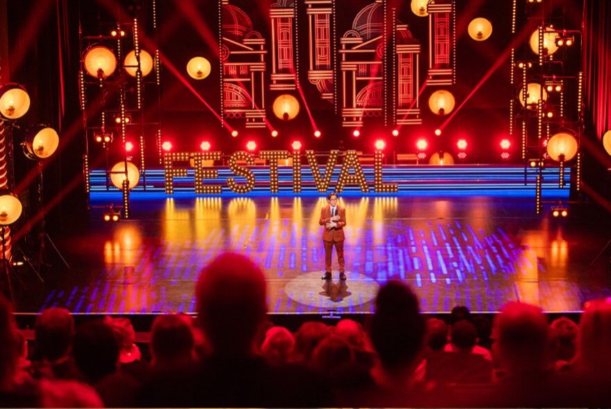View from the audience of a solo comedian performing on a large stage with a simple stencil graphic of Melbourne city as the backdrop.