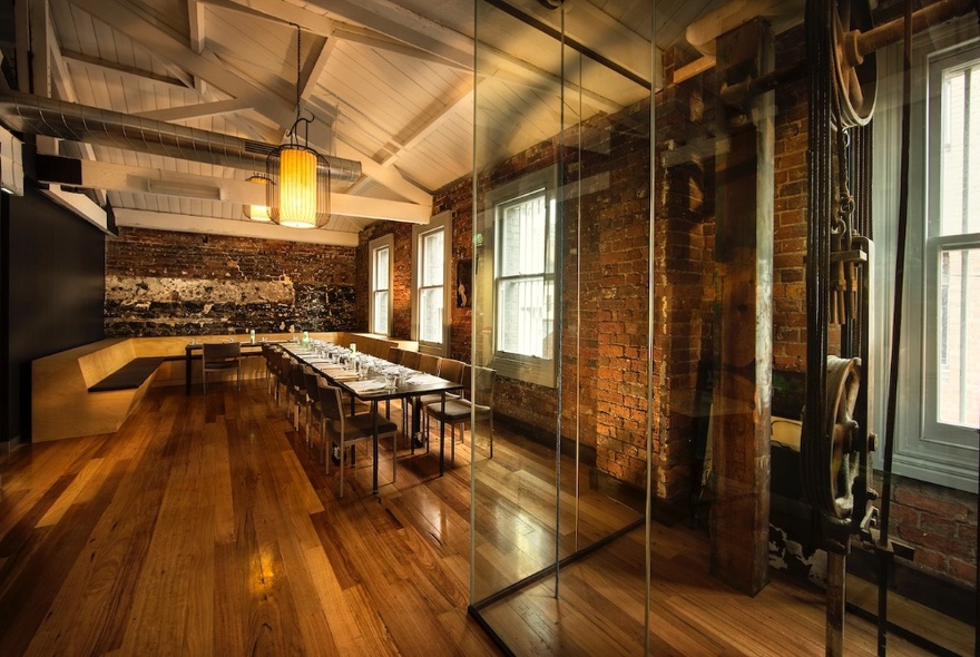 The interior of B'Churrasco, showing exposed brick wall, wooden floors, dining tables and chairs, hanging lights and a white wood panelled ceiling.