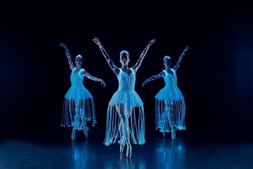 Three ballerinas on a darkened stage in glowing tutu dresses.