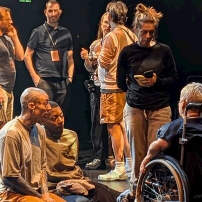A group of people standing around and talking on a darkened stage, with others sitting and talking to a person in a wheelchair.