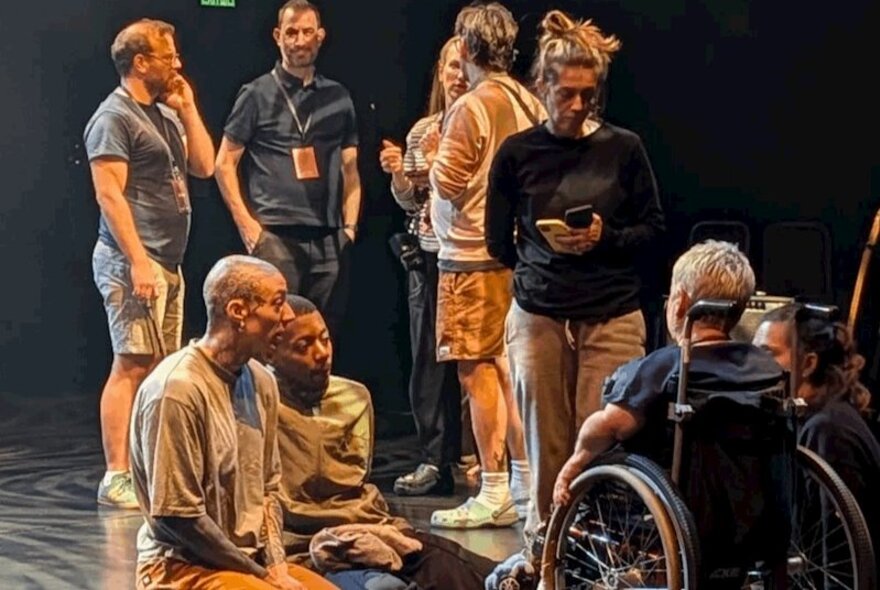A group of people standing around and talking on a darkened stage, with others sitting and talking to a person in a wheelchair.