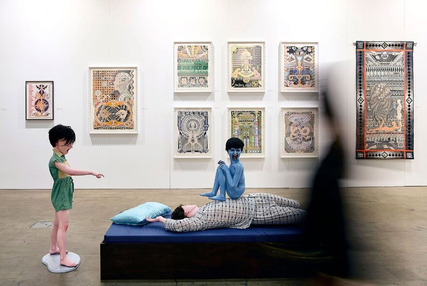 Art gallery with artworks and installations of children and adult reclining on a blue platform.