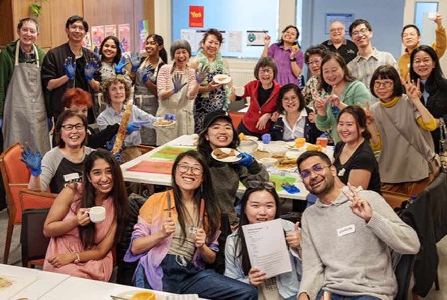 A smiling group of people seated around a table, some of them holding cups, others making a V peace sign with their fingers.
