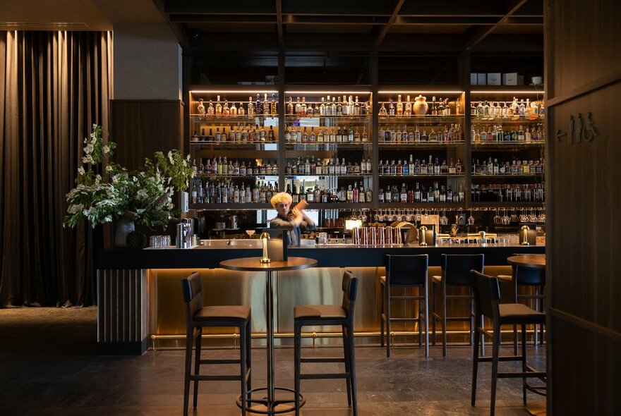 The bar at Next Hotel showing a bartender in front of a wall of bottled drinks, mixing a cocktail, with high tables and bar stools in the foreground. 