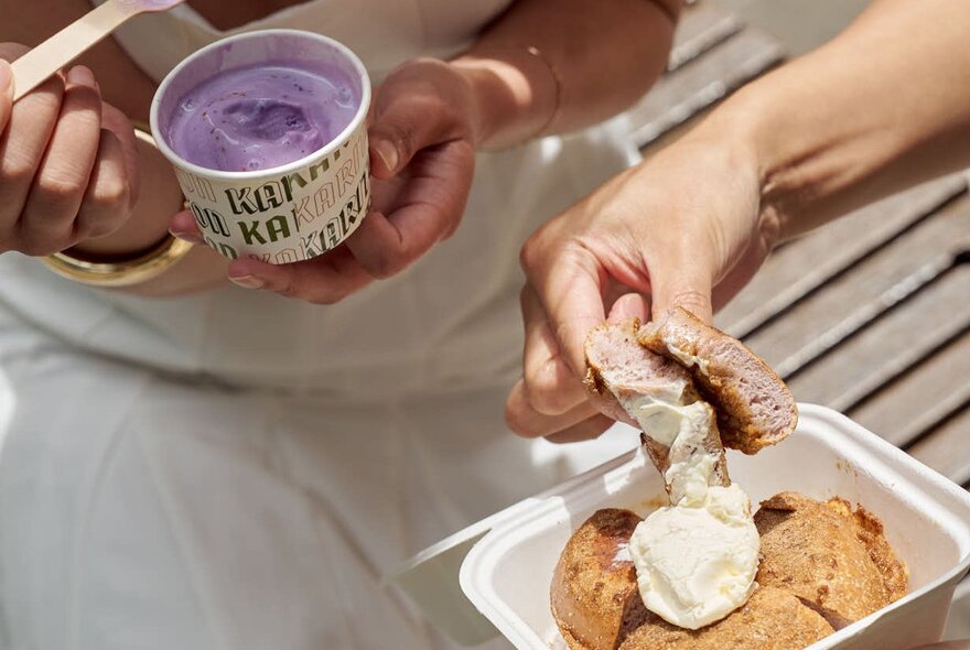 Two people enjoying a dessert outdoors, one person scoops purple ice cream from a cup and the other is dipping a bagel into cream.