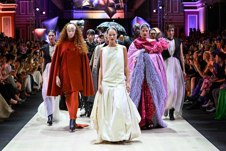 Fashion models walking down a catwalk runway with a seated audience watching, taking photos with their smartphones and applauding.