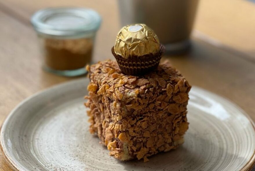 A small flake covered treat on a plate topped with a Ferrero Rocher gold foil covered chocolate.