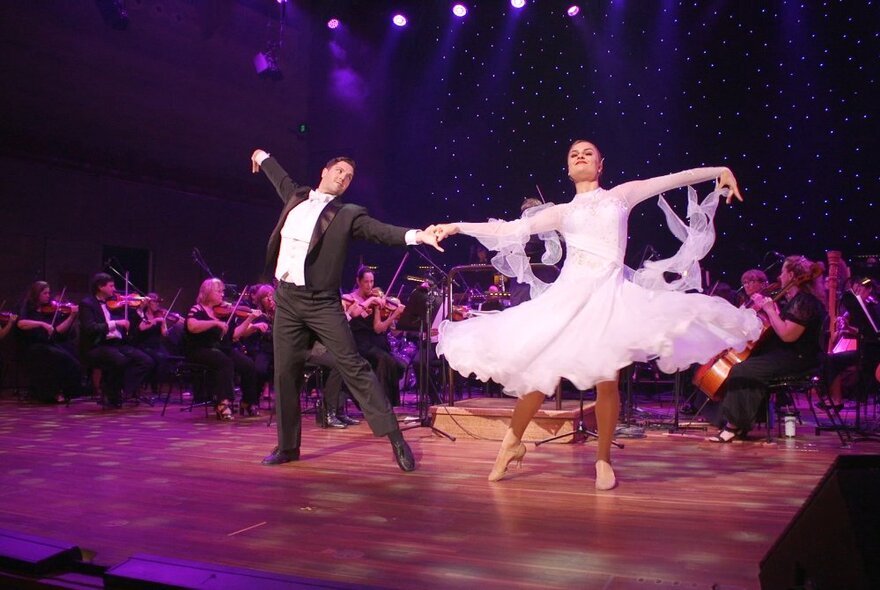 Two dancers twirling on stage in front of a large seated orchestra, the man wearing a tuxedo and the woman wearing a pink flouncy gown.