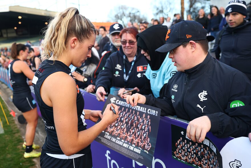 A Carlton Blues AFLW player signing a team poster for a supporter after a match.