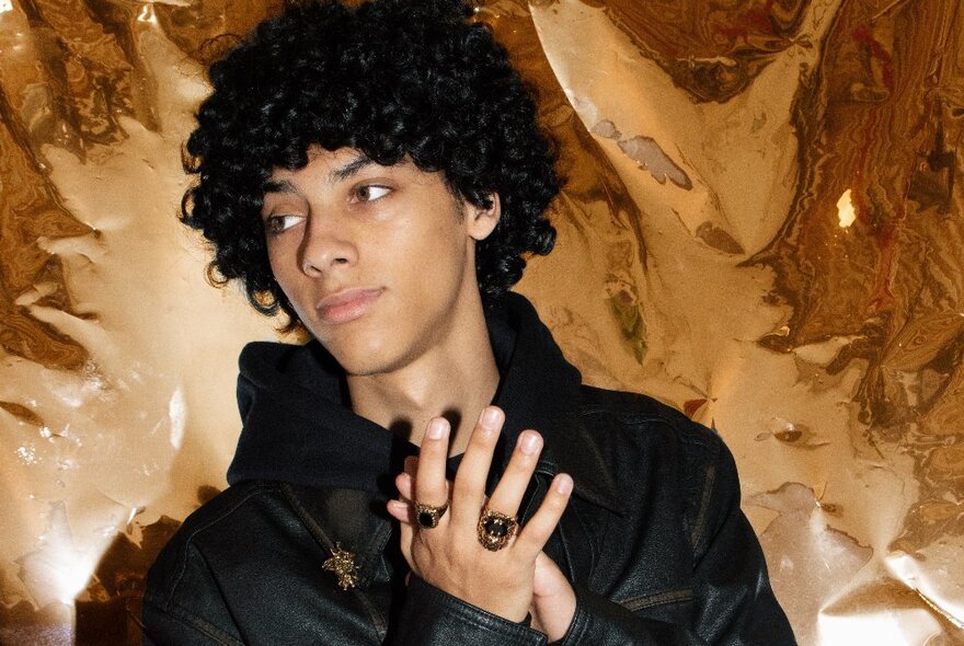 Male with curly hair wearing a dark top against a beige background.