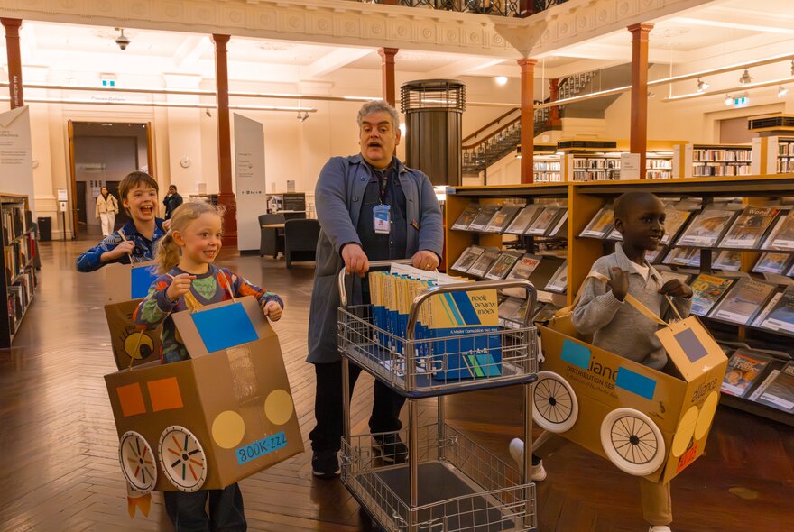 Three young children wearing cardboard box car creations in a library, with a librarian pushing a trolley.