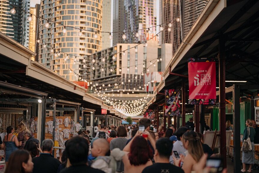 People walking around the Queen Victoria Summer Night market, with festive lights above them and a view of city buildings in the background.