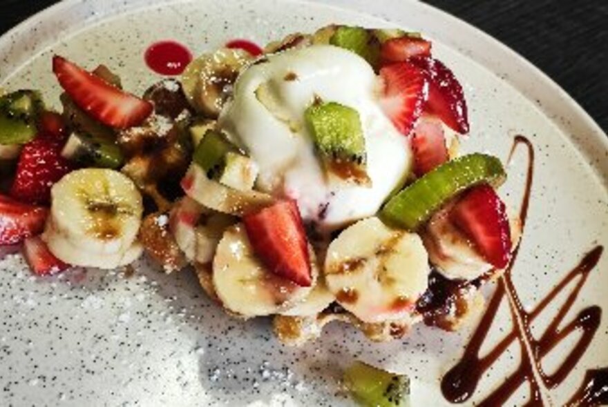 Chopped fruit and ice cream with chocolate sauce on a speckled plate.