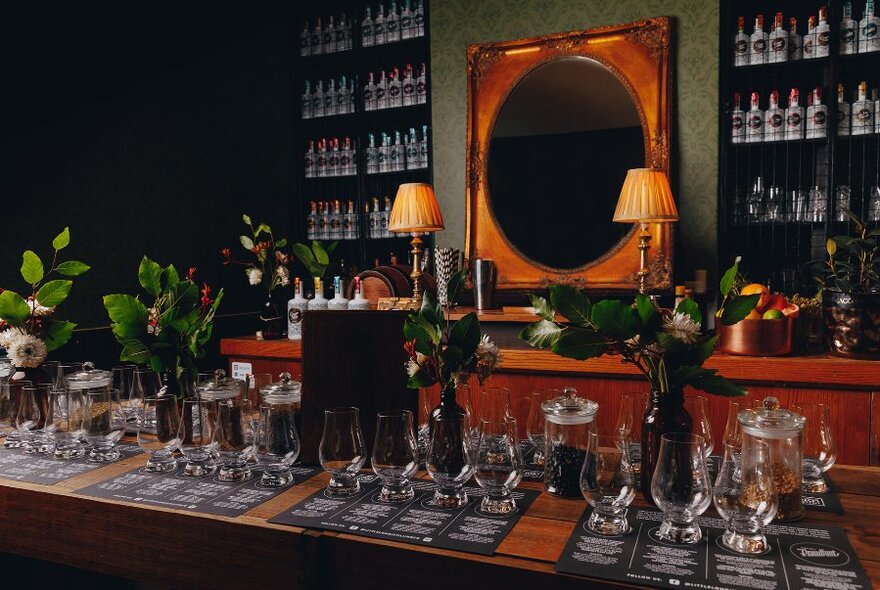 Interior of bar at Little Lon Distilling, with glasses set across the bar, a large bronze-framed mirror and lamps behind.