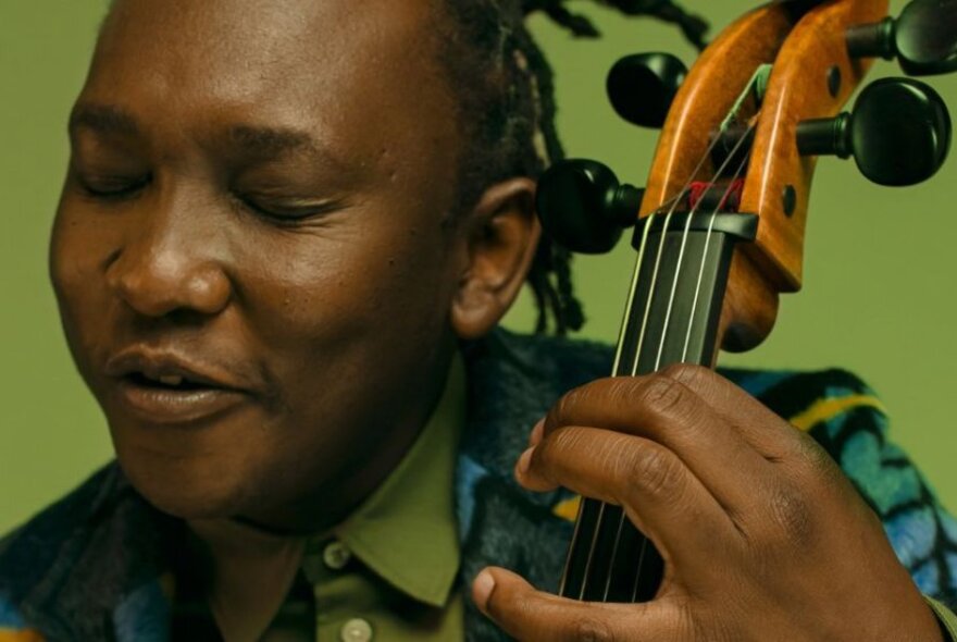 African musician Abel Selaocoe playing the cello, with dreads and colourful jacket.