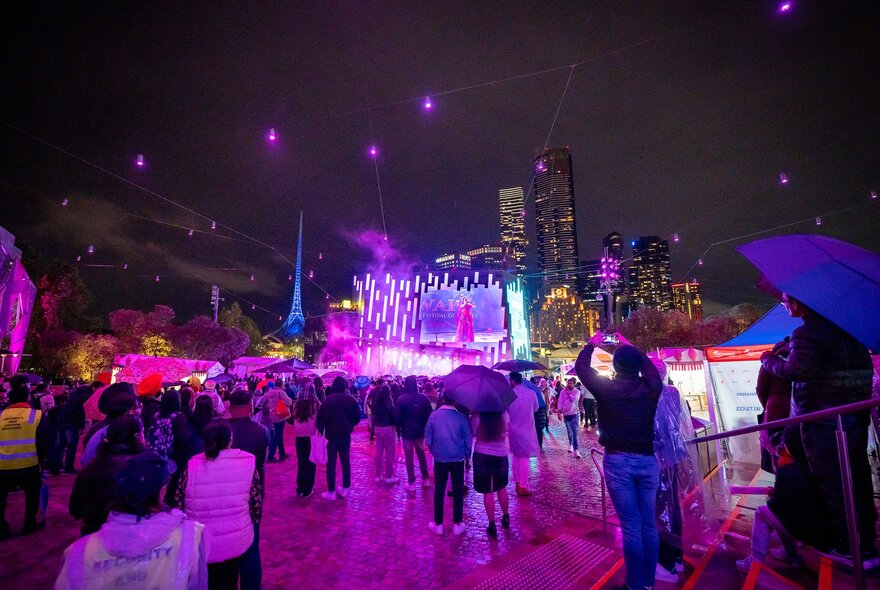 A crowd are gathered at night at Fed Square. They are watching a traditional Indian dance performance on the big screen. 