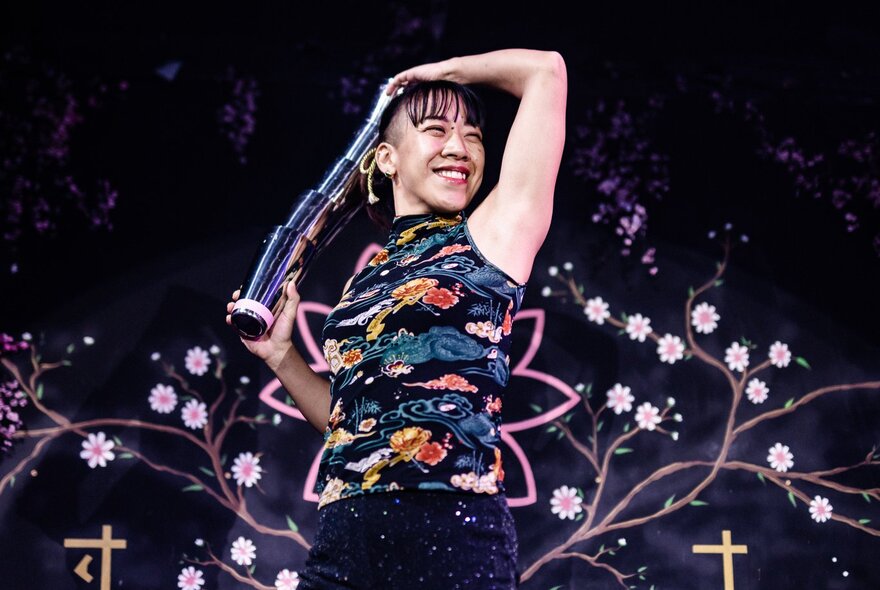 Magic performer wearing black and embroidered top, against an embroidered night sky.