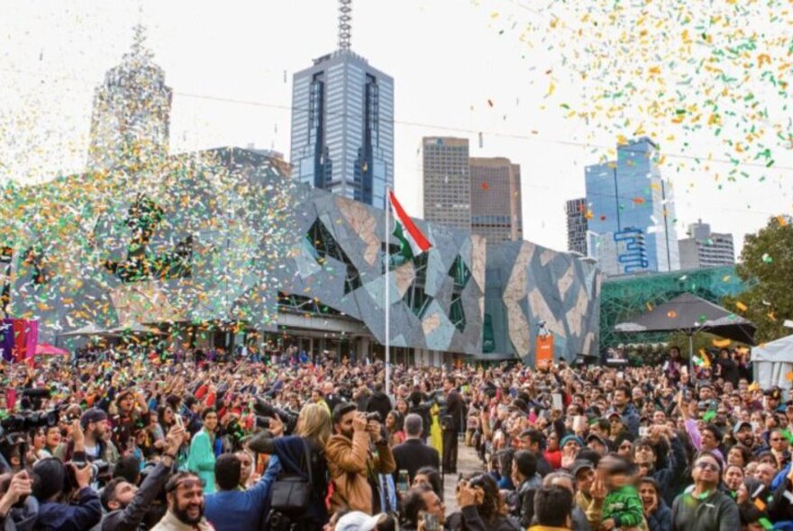 Orange, green and white bits of paper floating from the sky above a large crowd of people in the outdoor area at Fed Square, celebrating a flag raising ceremony.
