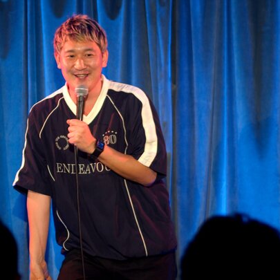 A stand-up comedian talking into a microphone they hold in their hand, performing live on stage.