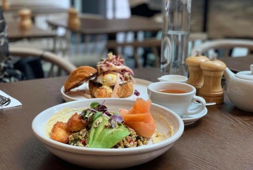 Avocado and salmon dish on a table, in front of an open, filled croissant and coffee.