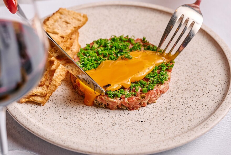 A knife and fork about to cut into a small, delicate tart wth chives and an orange sauce. 