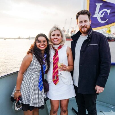Three people smiling for a photograph on the bow of a river cruise vessel with the Port of Melbourne in the background.