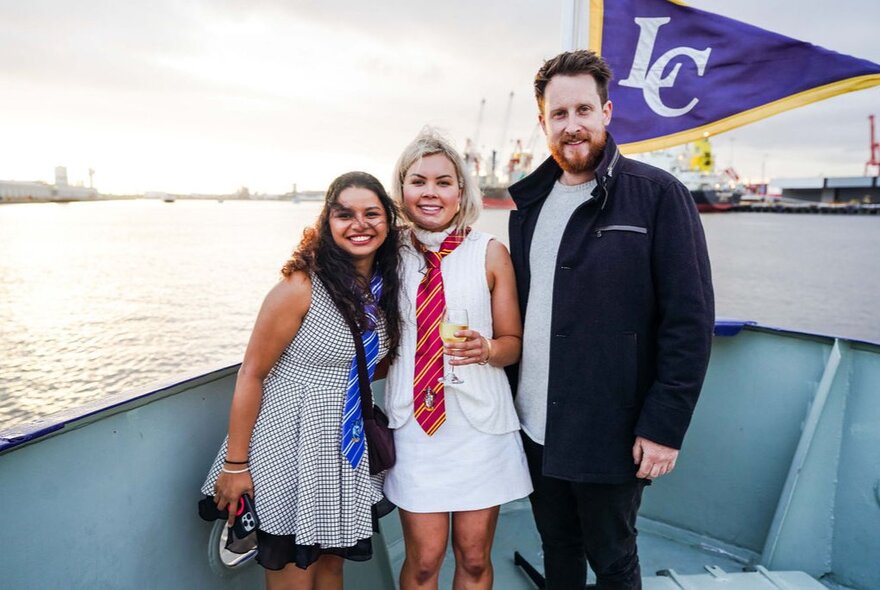 Three people smiling for a photograph on the bow of a river cruise vessel with the Port of Melbourne in the background.