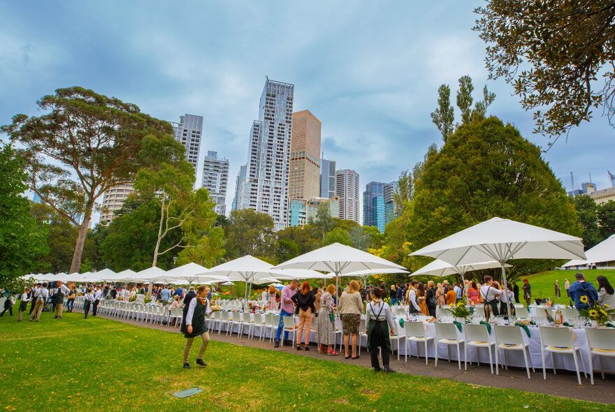 Outdoor dining event in a park with long tables and white umbrellas with trees and city buildings in the background.