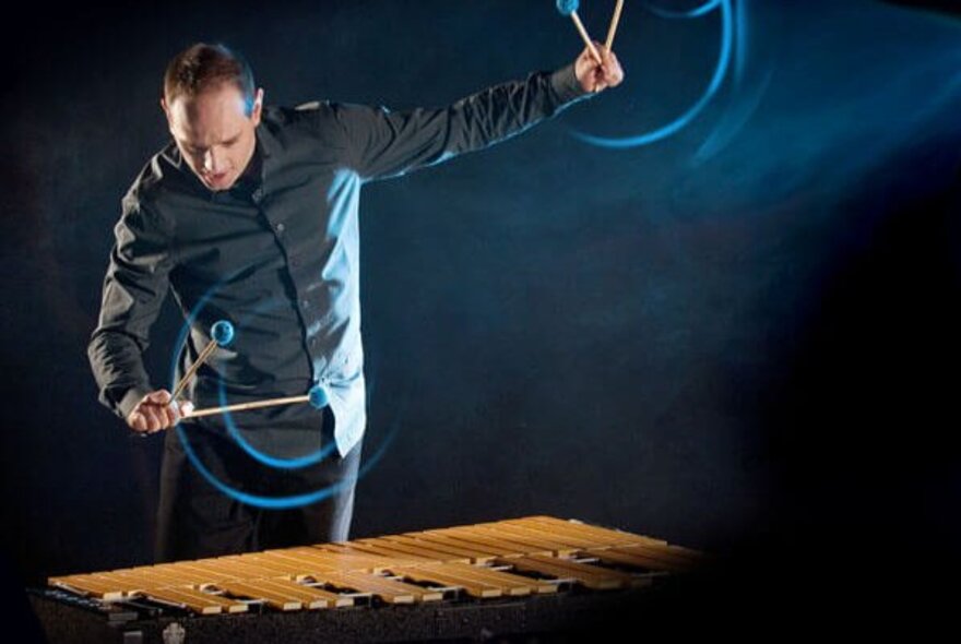 A man playing a vibraphone with two sticks in each hand, one arm raised above his head. 