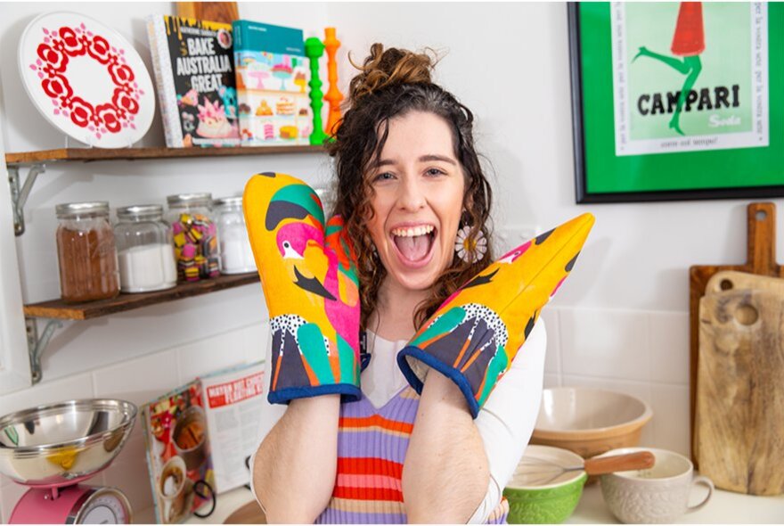 A person holding up colourful, bird-printed cotton oven mitts with a bright yellow and multi-coloured pattern.