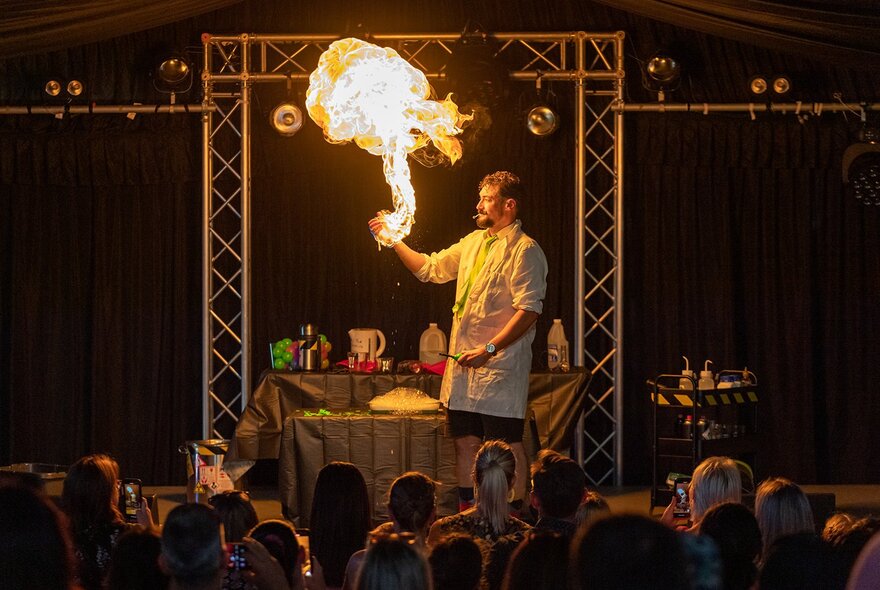 A man in a white lab coat on stage with a large flame coming from his raised hand with an audience looking on. 