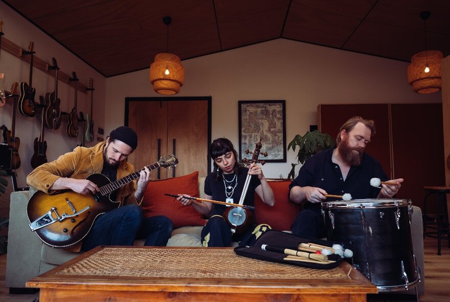 Three musicians playing traditional and Western instruments in a room with instruments and soft lighting.