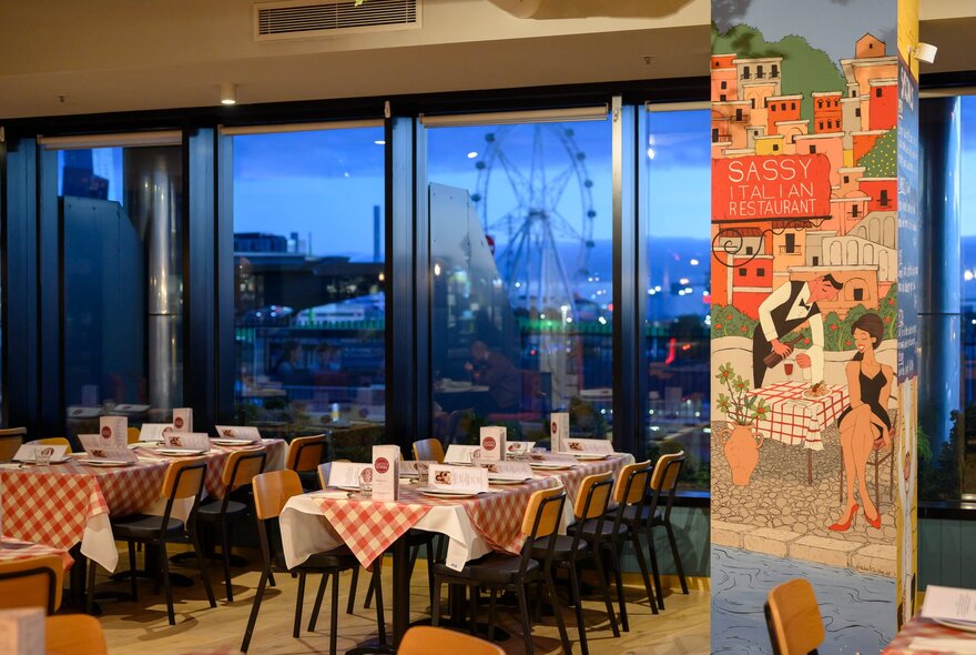 Interior of Sassy Italian Restaurant, with long tables covered in red and white checked tablecloths, with chairs, set for service and large windows in the background with views of Docklands.