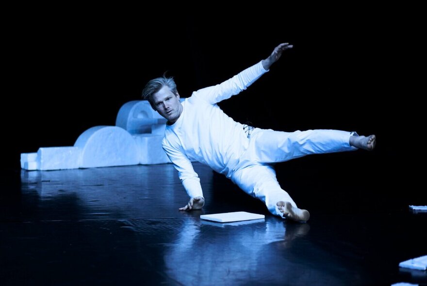 A dancer wearing all white performing a dance on a theatre stage floor, with one hand and one leg resting on the ground.