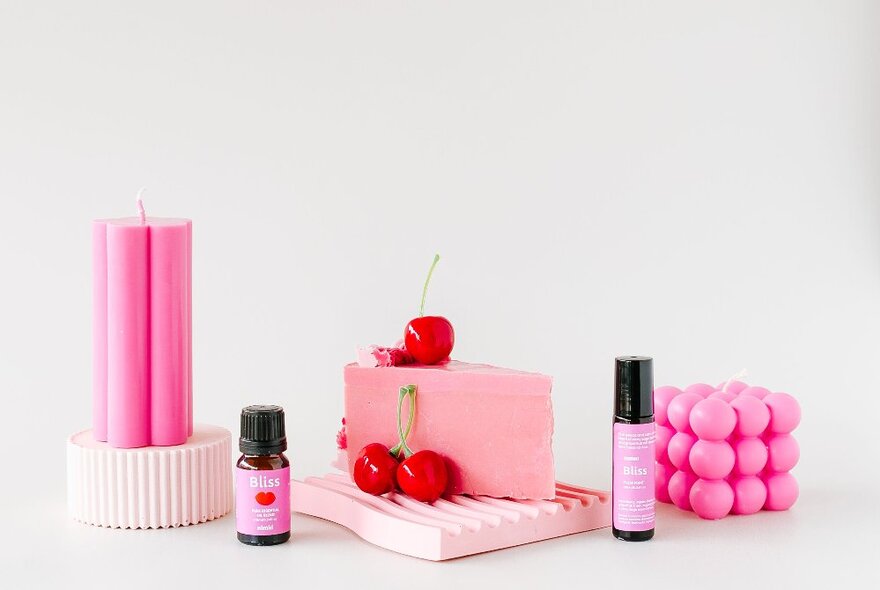 A collection of pink soaps, candles and serums with three bright red cherries.