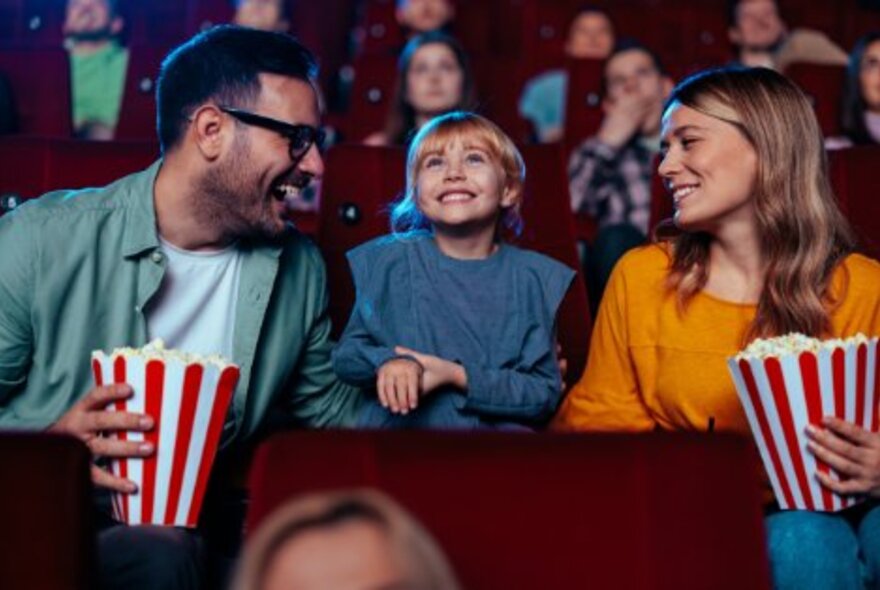 Parents and child seated in cinema seats, parents holding large striped containers of popcorn, the child in the middle smiling up at the screen.