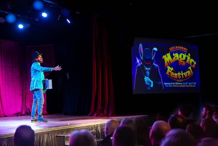 Magician performing on stage, wearing a pale blue jacket with an arm outstretched.