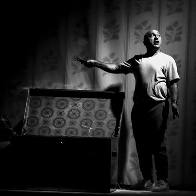 The actor George Kapiniaris standing next to an open chest or glory box under a spotlight on a theatre stage.