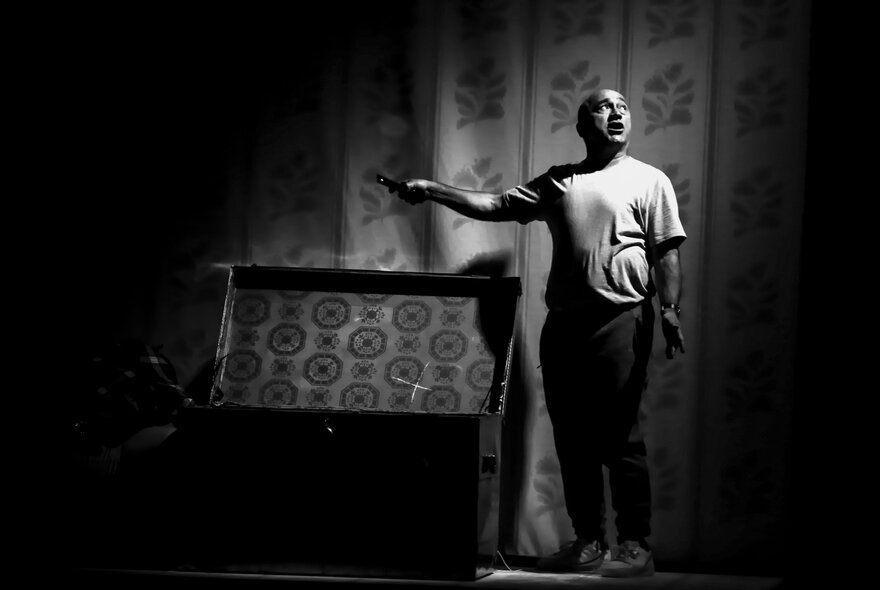 The actor George Kapiniaris standing next to an open chest or glory box under a spotlight on a theatre stage.