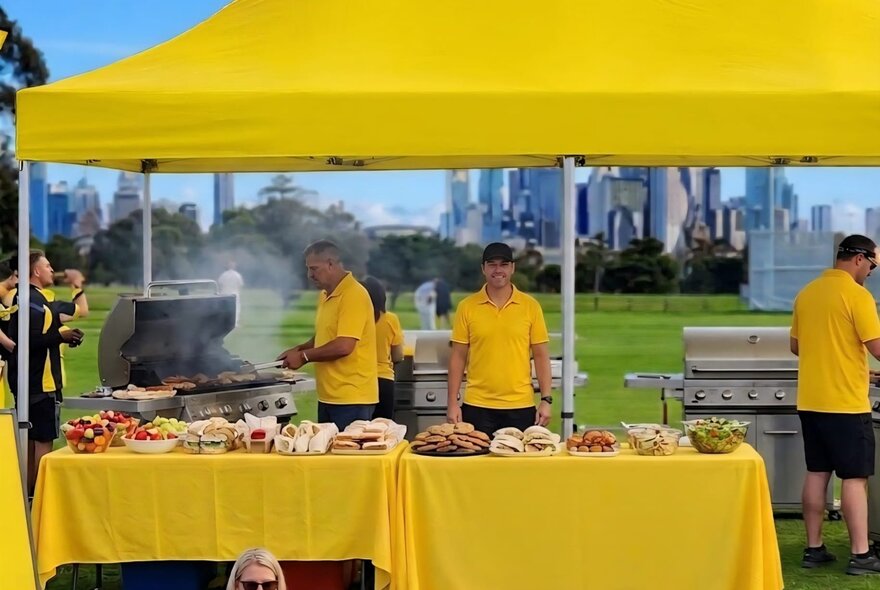 An outdoor barbeque event in a park, with people wearing yellow t-shirts working under a yellow shade umbrella and behind yellow tables managing a large food booth.
