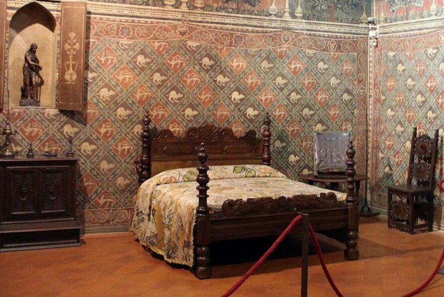 Early Renaissance bed with dark wood head and base, with patterned wallpaper and statue in a niche.