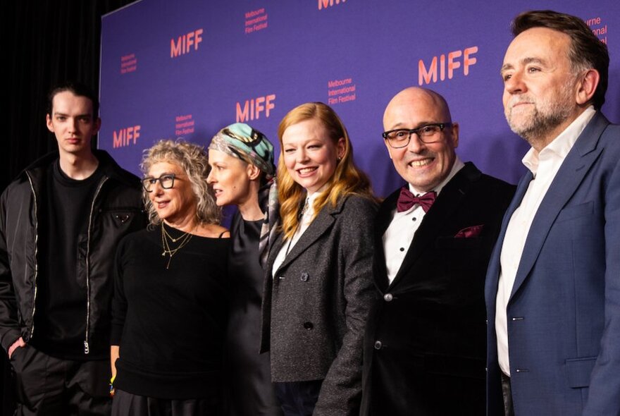 A group of six actors and actresses standing in a line in front of a purple branding board with MIFF printed on it. 