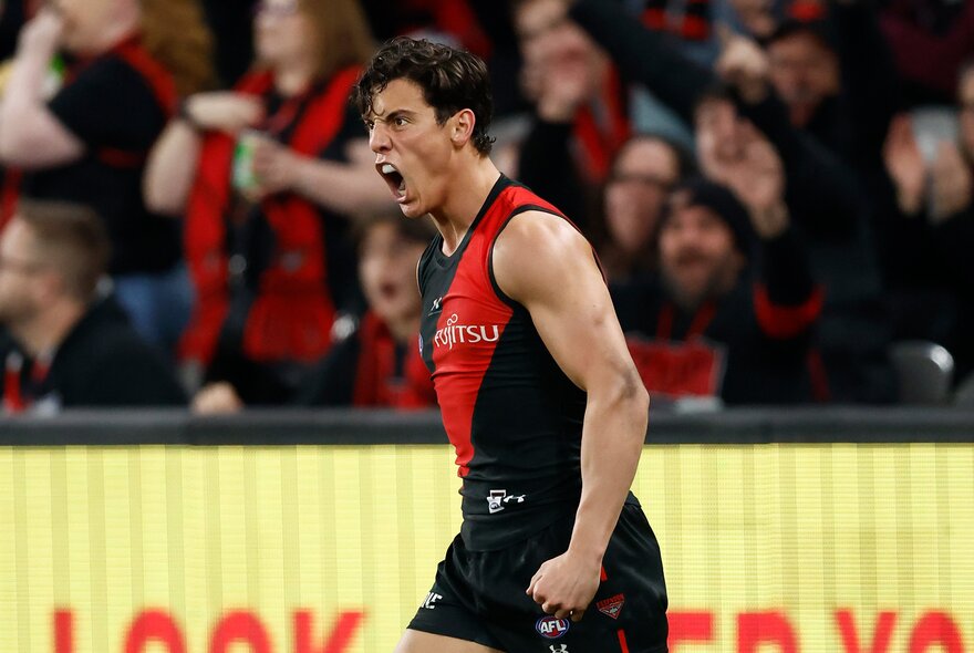 An Essendon AFL football player with pumped fists during a match, with the blurred spectators in the stands behind him.