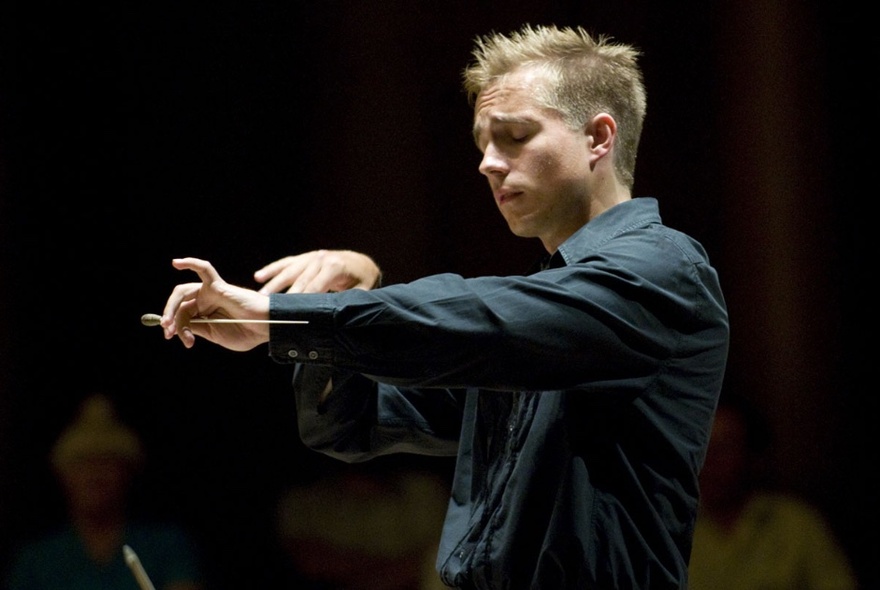 An orchestra conductor in a black shirt, arms outstretched and eyes closed, conducting a performance.